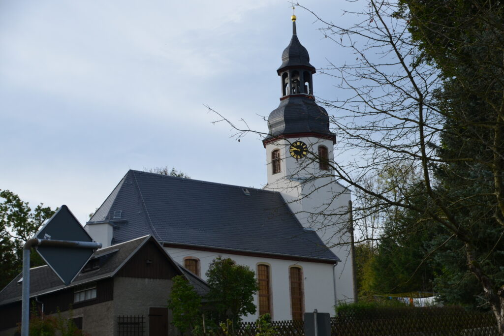 Weiße Dorfkirche mit barockem Turm. Davor stehen Bäume und ein Wohnhaus.