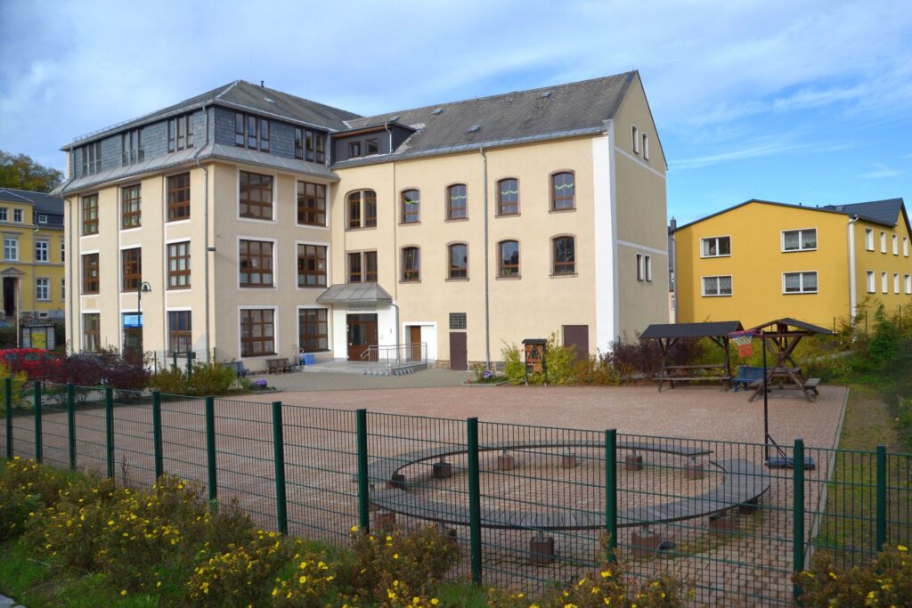 Großes Schulgebäude mit Schulhof. Davor stehen Spielgeräte und eine Sitzgruppe.