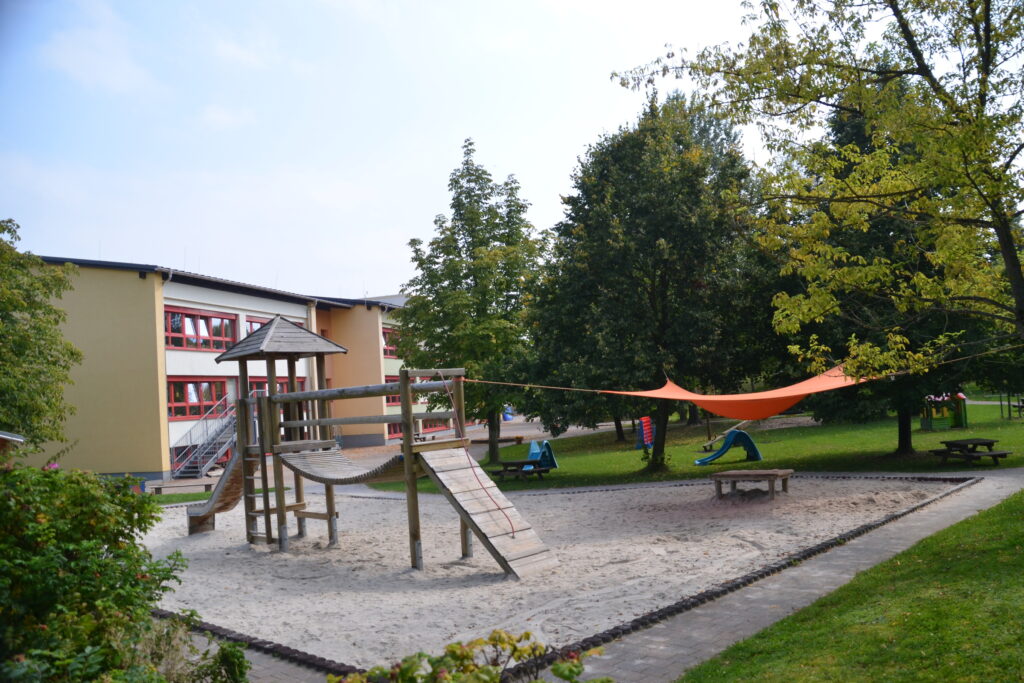 Kita-Gebäude mit großem Spielplatz. Davor stehen ein Klettergerüst und ein Sonnensegel.