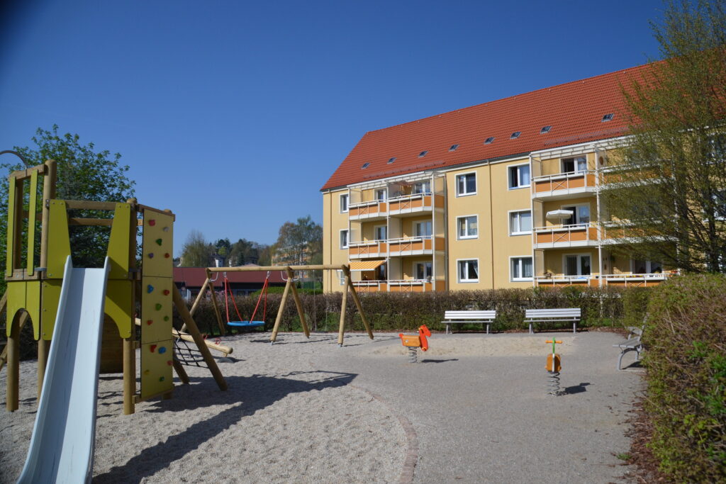 Spielplatz mit Rutsche, Schaukel und Federwippe. Im Hintergrund ein Wohnhaus.